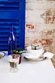 A glass of white wine on a table with a white tablecloth, stacked crockery, kalamata olives and a wine bottle in front of a royal blue wooden shutter