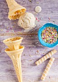 A melting scoop of vanilla ice cream surrounded by cones and colourful sugar sprinkles on a purple wooden surface