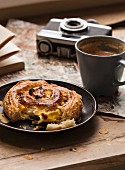 A cinnamon bun with raisins, a cup of coffee, a camera and a map on the wooden table
