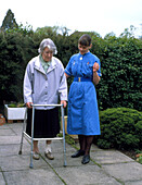 District nurse helps old woman use walking frame