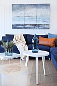 White side tables and dark blue couch below seascape painting on white wooden wall