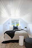 Doppelbett mit heller und dunkler Tagesdecke vor Fenster in Dachzimmer mit weisser Holzverkleidung