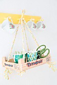 Seedlings in paper cups in recycled wooden crate hung from yellow coat rack by ribbons