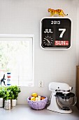 Küchenkräuter, Obstkörbchen und Küchenmaschine, darüber Kalenderuhr an weisser Fliesenwand