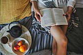Frau liegt lesend auf Bett, daneben Tablett mit Tee und Obst