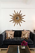 Scatter cushions with stag motifs on black velvet sofa below sunburst mirror on white wall