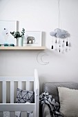 Kinderzimmer mit weißem Gitterbett und grauem Sessel, darüber Ablage und Mobile mit Wolkenmotiv