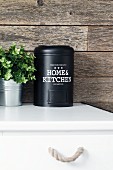 Black metal tin and foliage plant on kitchen cabinet against rustic wood cladding