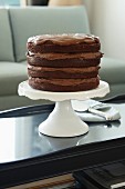 A multilayered chocolate cake on a cake stand on a coffee table