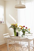 Schlichter, weisser Esstisch mit bunten Blumensträussen und Klappstühlen vor Fenster