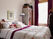 Double bed with ethnic bedspread, scatter cushions and white bed linen with pompoms next to wardrobe