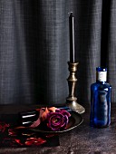Dark purple rose on dish, blue glass bottle and brass candlestick in front of black curtain