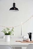 Vase of ranunculus and beaker of black drinking straws on white dining table in front of pink garland on wall