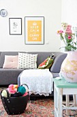 White crocheted blanket and various cushions on grey sofa below black-framed pictures