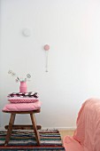 Stack of pink cushions on rustic wooden stool on rag rug and necklace hanging from wall peg in bedroom