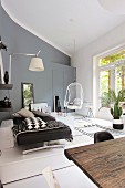 Couch, hanging chair, wall lamp and plants in modern living room in shades of grey with rustic wooden table in foreground