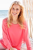 A young blonde woman sitting on a beach wearing a long-sleeved, layer-look top