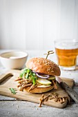 Puled pork burger with apple sauce, gherkins, onions and apple slices on a chopping board with a glass of beer in the background