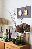 Branches of blossom and hydrangeas in glass vases next to wine bottles on sideboard