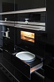 A kitchen cupboard with a shiny black surface, a built-in oven and plates in an open warming drawer