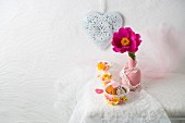 A cupcake decorated with paper flowers in front of a peoney in a vase and a decorative heart