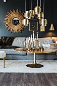 Candlesticks on round coffee table below brass pendant lamps and sunburst mirror on blue wall