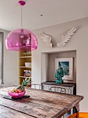 Rustikaler Eisentisch mit Holzplatte und pinkfarbene Pendelleuchte vor Wandnische mit Anrichte und Kunstobjekten