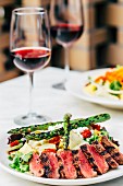 Grilled beef steak with asparagus salad and glasses of red wine on a table in a restaurant