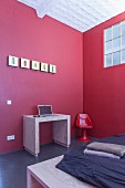 Laptop desk against red wall in minimalist workspace in corner of bedroom with industrial windows
