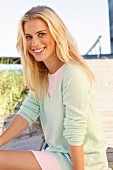 A young blonde woman on a beach wearing a pastel green jumper and a pink mini skirt