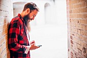A man with a beard and a lumberjack shirt leaning on a wall with his smartphone in his hand