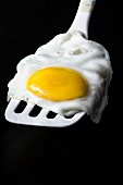 A fried egg on a white spatula on a black background