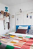 Schlafzimmer mit bunter Strick-Patchworkdecke, Betthaupt aus Holzbrettern und Kleidungsstücken an Hakenleiste