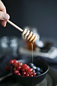 Honey dripping from a dipper onto berries in a dark bowl