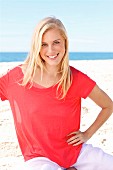 A young blonde woman sitting on a beach wearing a red top