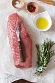 A raw beef steak with spices, oil and rosemary on paper