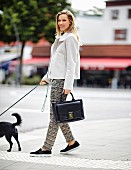 Blonde Frau in Wolljacke, gemusterter Stoffhose, Sneaker und Henkeltasche geht mit Hund spazieren
