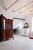 Antiker, kunsthandwerklicher Holzschrank in weißem Dachzimmer mit Kronleuchter und nostalgischem Flair