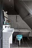 Free-standing bathtub against mosaic-tiled wall below grey sloping ceiling