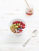 Oat granola crumble with fresh fruit, berres, honey in bowl over white painted wooden background