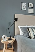 Black lamp on bedside table next to bed with beige headboard and grey cover against grey wall