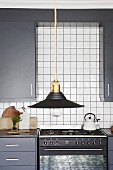 Schwarzer Lampenschirm vor grauer Einbauküche mit weißen Wandfliesen und Gasherd