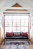 Couch with scatter cushions in bright gable window with chipboard cladding and recycled board floor