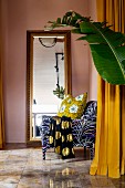 Patterned armchair in front of full-length mirror with large leaf in foreground