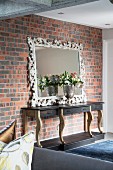 Artistic mirror leant against brick wall and flowers in silver goblet on top of console table