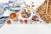 Oat granola with nuts, yogurt, honey, fresh figs and blueberries in bowl on white wooden background