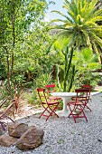 Weisser Tisch mit roten Stühlen auf Kiesfläche vor exotischen Pflanzen