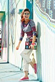 A brunette lady wearing beige-coloured chinos, a striped jumper, sunglasses, a hairband and sneakers leaning against a colourful exterior wall