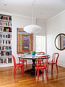 Dining area with a round table and red bentwood chairs in an old building with a bookcase and a colorful painting