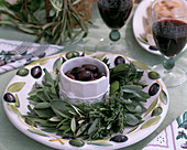 Plate wreath of sage, thyme, laurel and olive branches, bowl with black olives